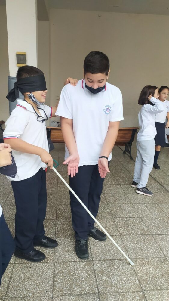 En la imagen se ven dos alumnos haciendo taller de accesibilidad, uno de ellos con bastón blanco y los ojos tapado, el otro lo guía. 