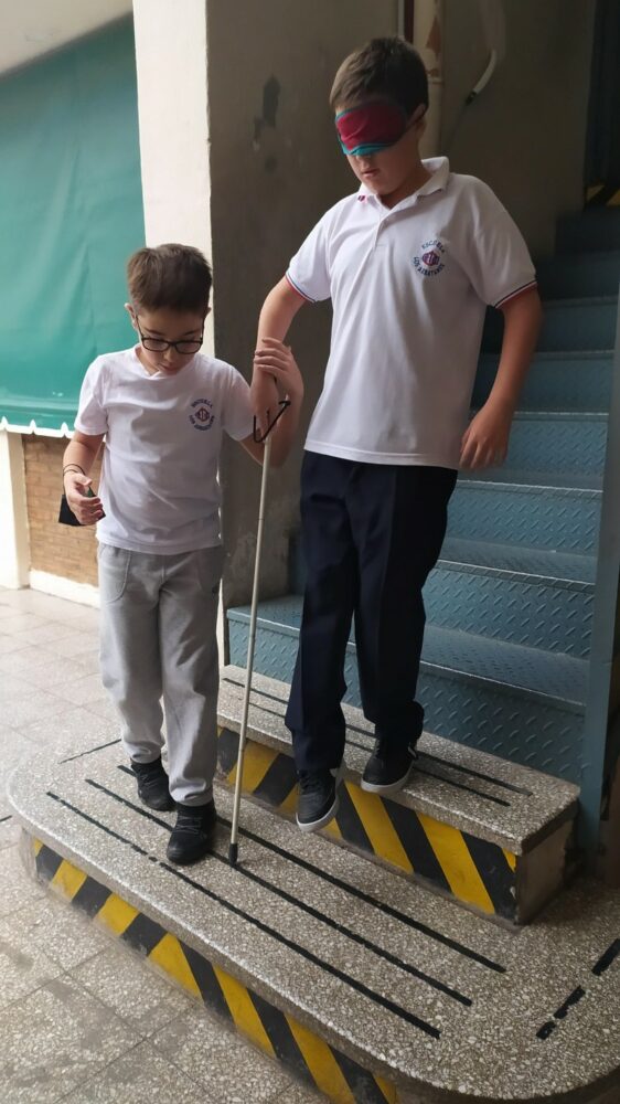 En la imagen se ven dos alumnos bajando una escalera, uno de ellos con bastón blanco y los ojos tapado, el otro lo guía. 